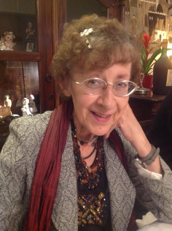Portrait de Jany, souriante, la joue appuyée sur sa main. Elle porte des lunettes fines, un gilet gris clair, une écharpe bordeaux et un collier de perles colorées.